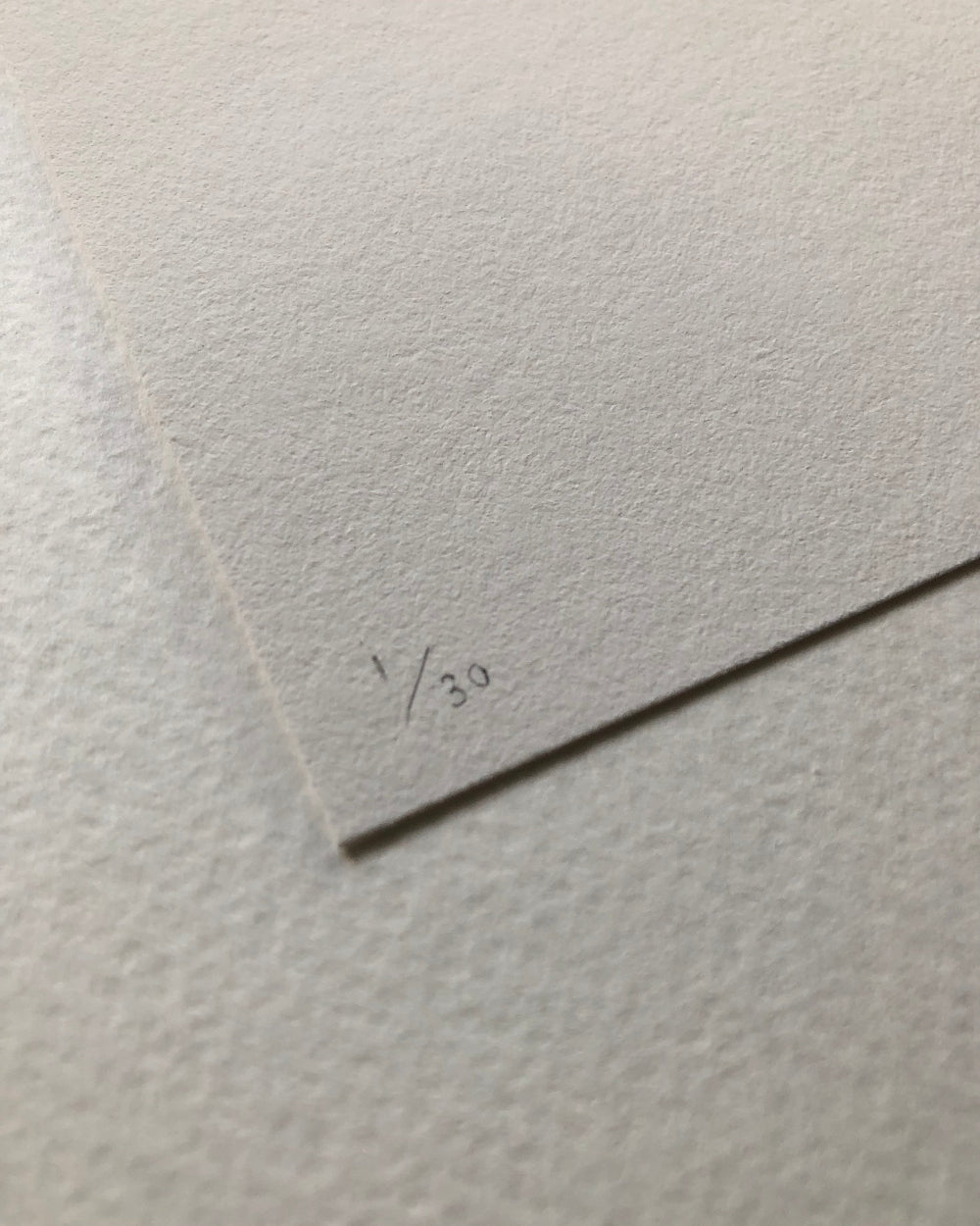 A close-up of textured white paper with  small hand writting reading 1/30 near the corner, photographed on a matching light surface.