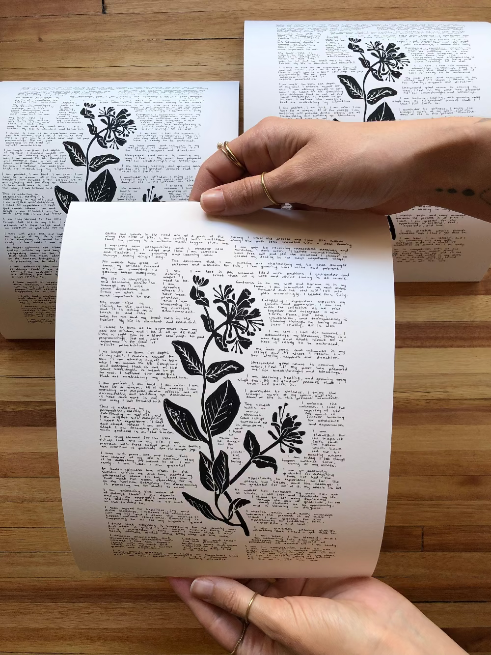 Three lino prints of a honeysuckle flower surrounded by tiny handwritten positive affirmations photographed together on a table. One is being held by a person for perspective.