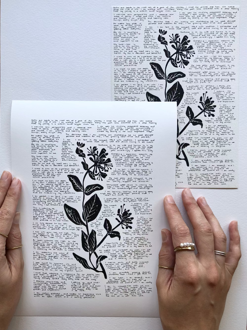 A lino print of a honeysuckle flower surrounded by tiny handwritten positive affirmations photographed flat on a table next to the original artwork.