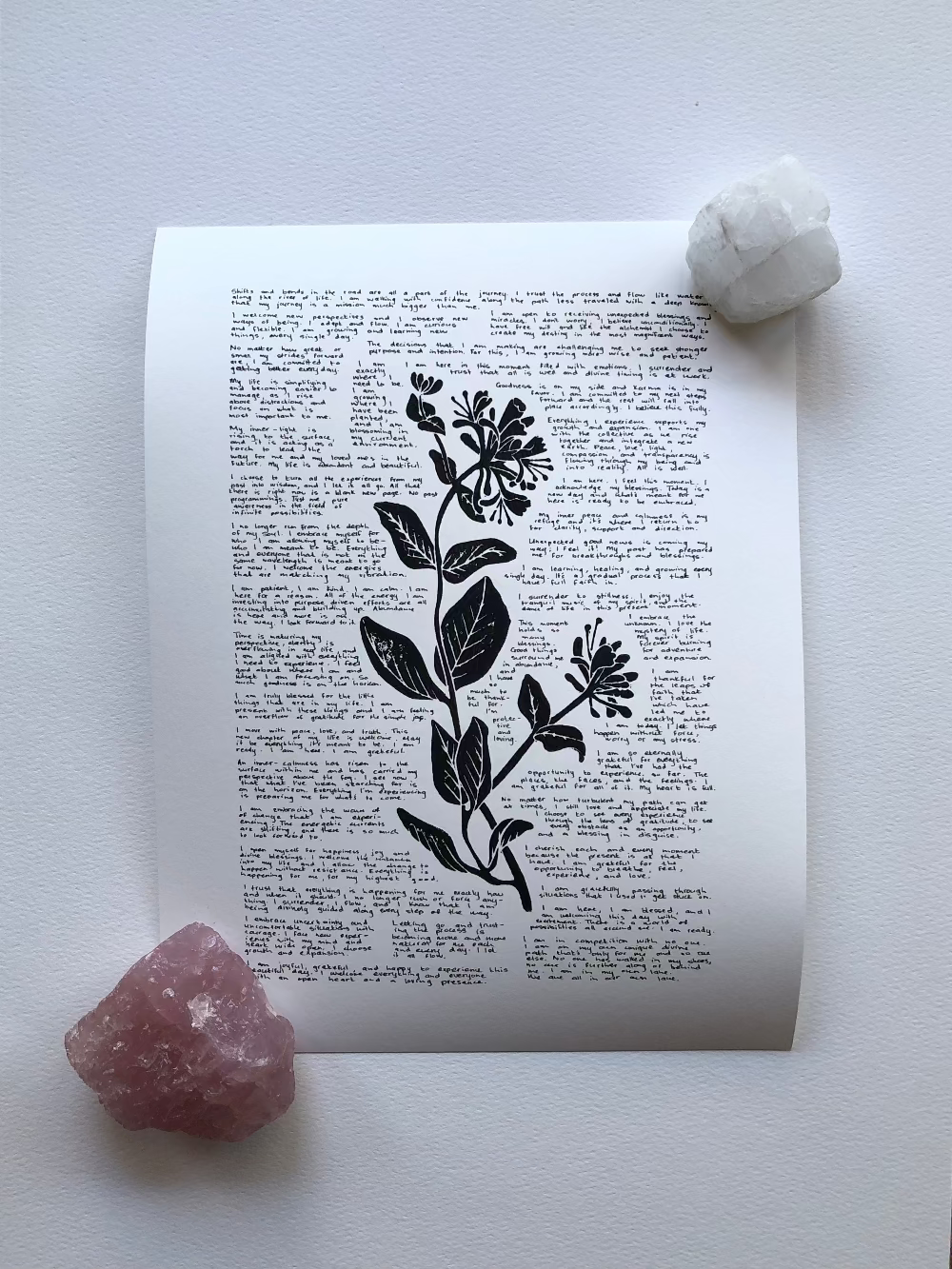 A lino print of a honeysuckle flower surrounded by tiny handwritten positive affirmations photographed flat on a table and held down with two crystals.
