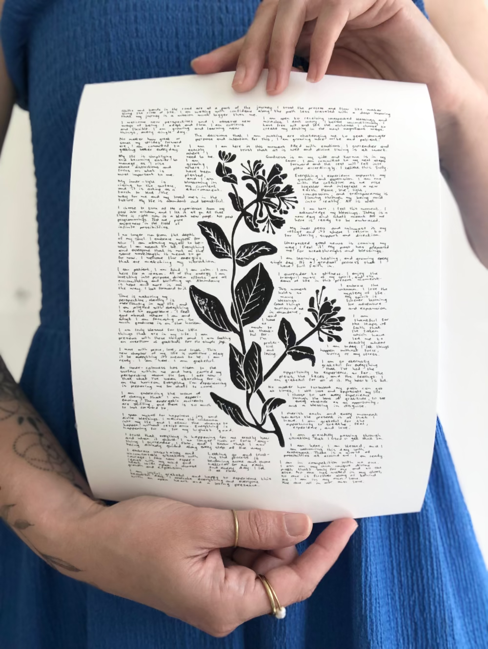 A lino print of a honeysuckle flower surrounded by tiny handwritten positive affirmations photographed being held by the artist.