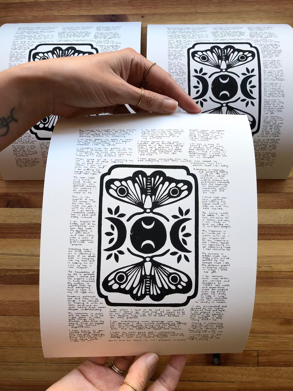 Three lino prints of moths and moons surrounded by tiny handwritten positive affirmations, photographed on the table. One is being held by a person to show perspective.