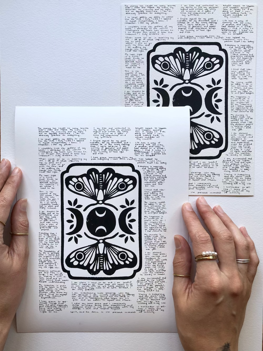 Lino print of moths and moons surrounded by tiny handwritten positive affirmations, photographed flat on a table next to the original artwork.