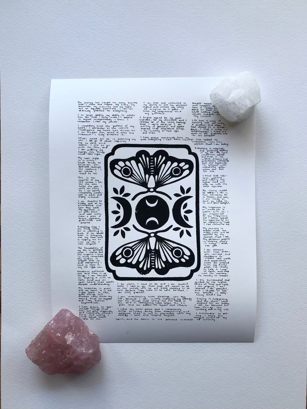 Lino print of moths and moons surrounded by tiny handwritten positive affirmations, photographed flat on a table held down by two crystals.