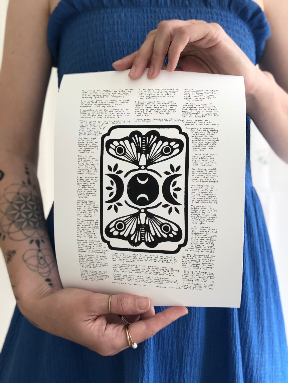 Lino print of moths and moons surrounded by tiny handwritten positive affirmations, photographed being held by the artist.