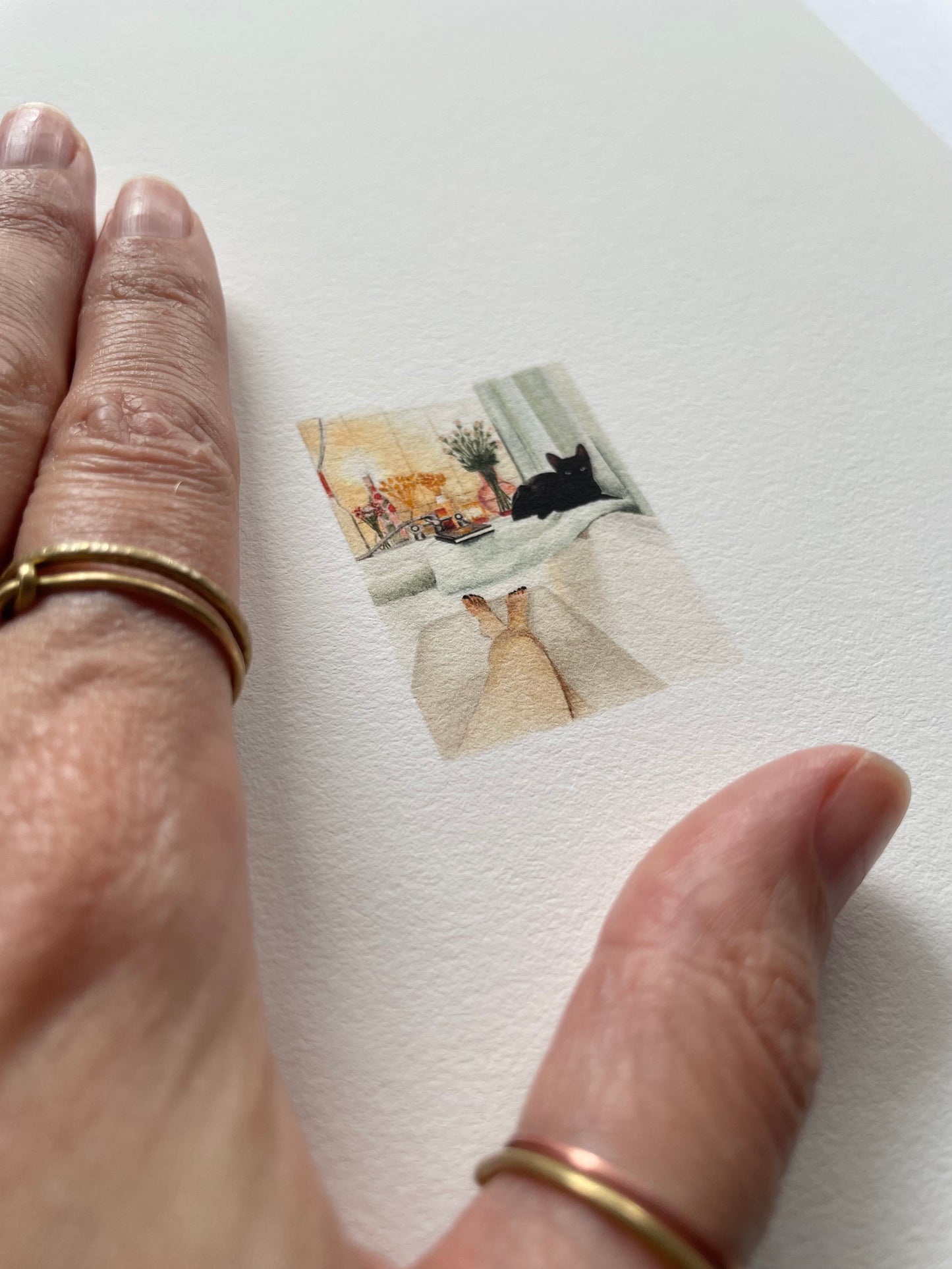 A hand resting on top of light textured paper with a tiny print of a cat lying by a lady while she baths