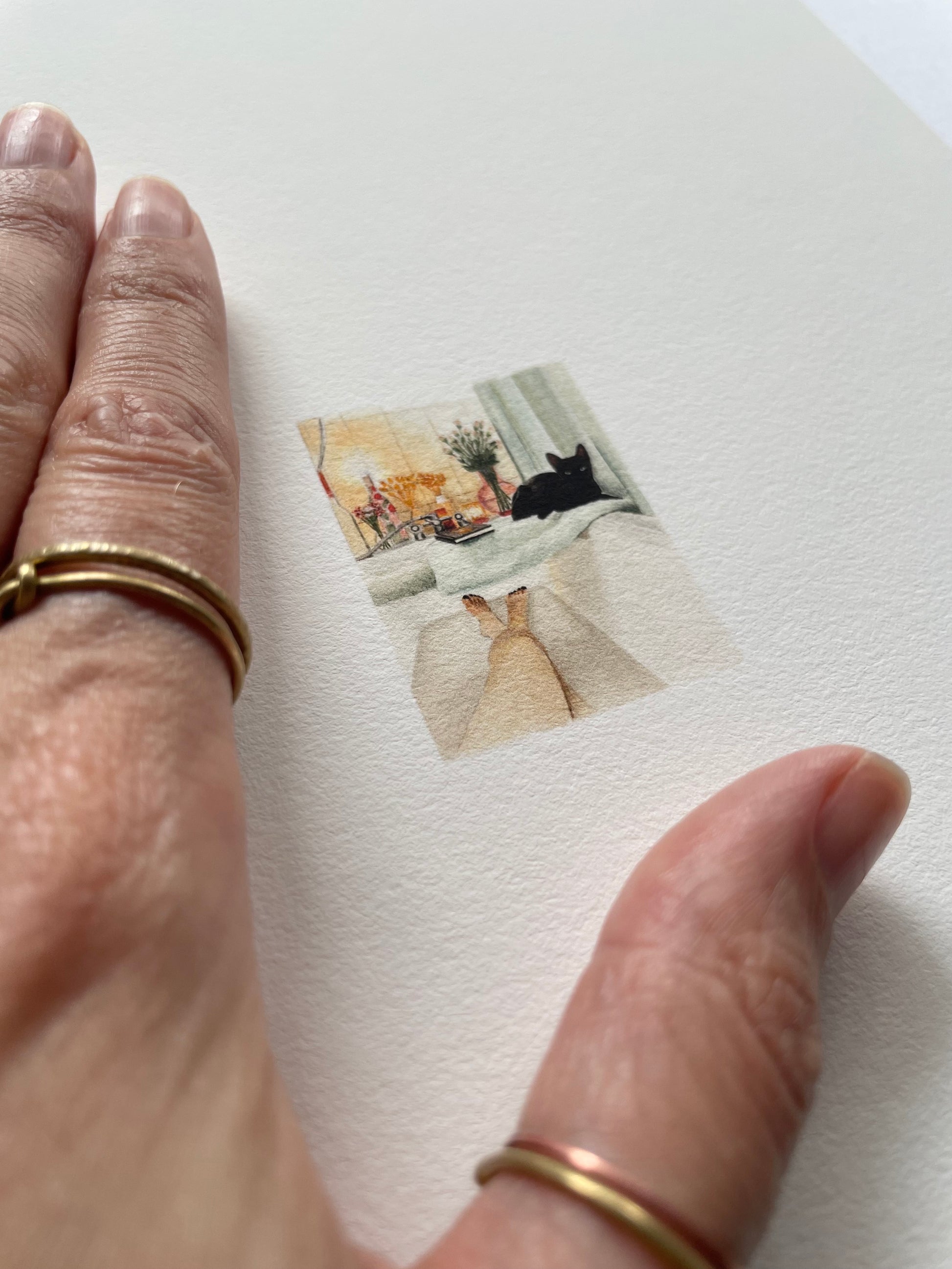 A hand resting on top of light textured paper with a tiny print of a cat lying by a lady while she baths