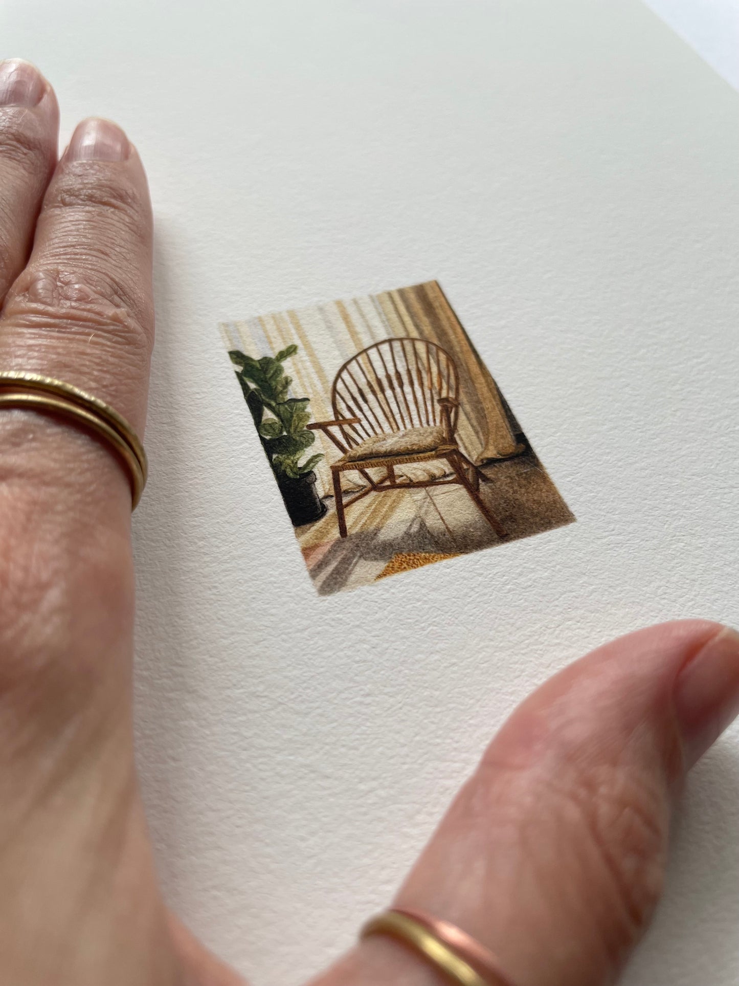 Hand resting on top of light textured paper with a tiny print of a wooden chair in a room with curtains, a plant and sun filtering through