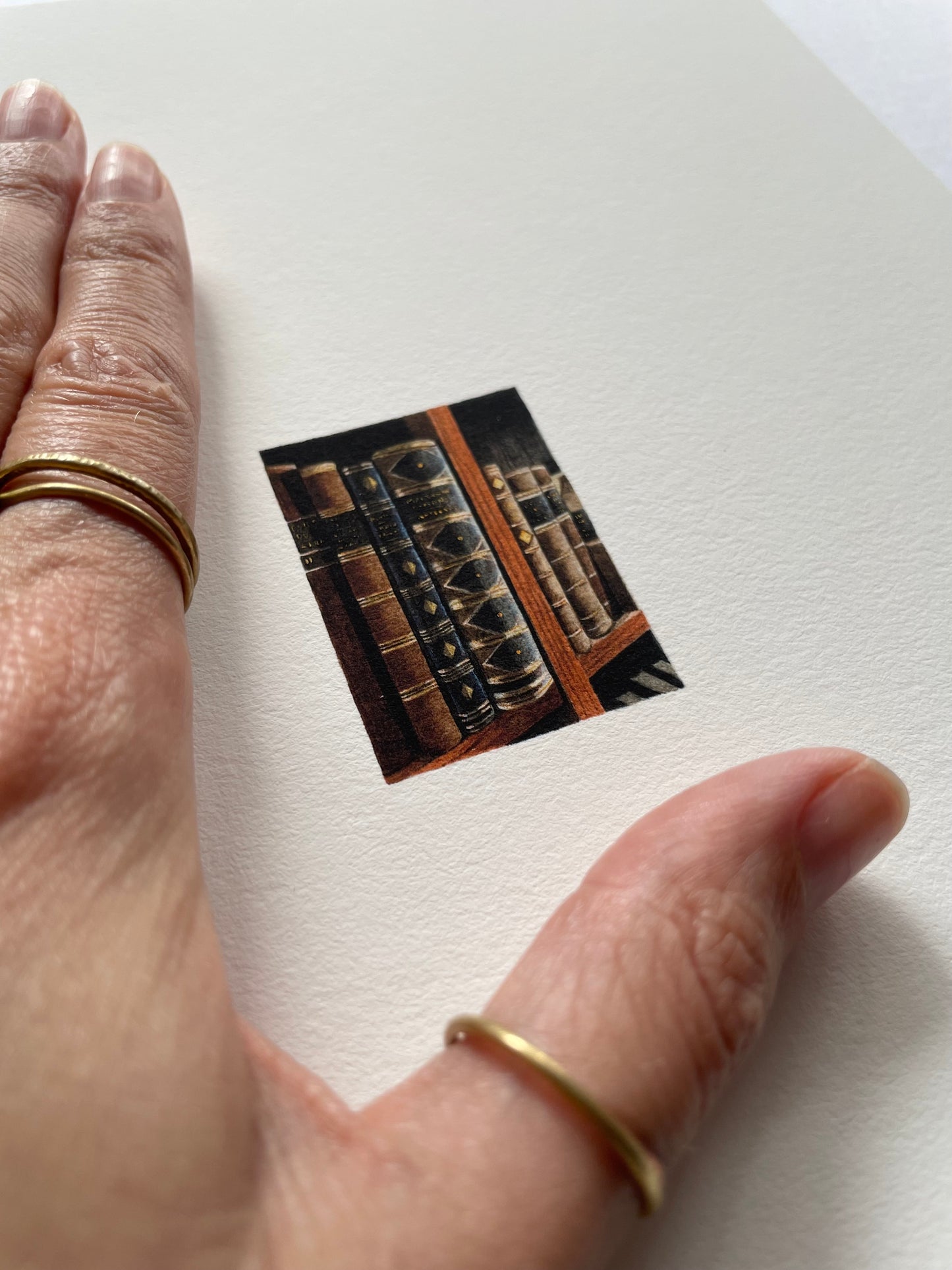 A hand resting on light textured paper of a tiny print of antique books on a bookshelf