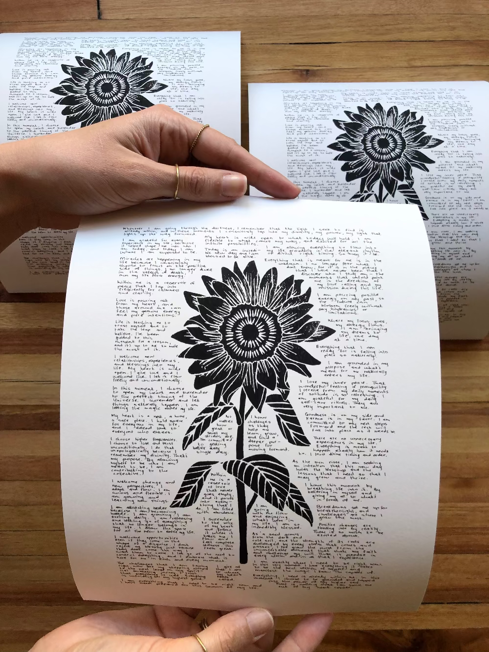 Three lino prints of a sunflower surrounded by tiny handwritten positive affirmations photographed together on a table. One is being held by a person for perspective.