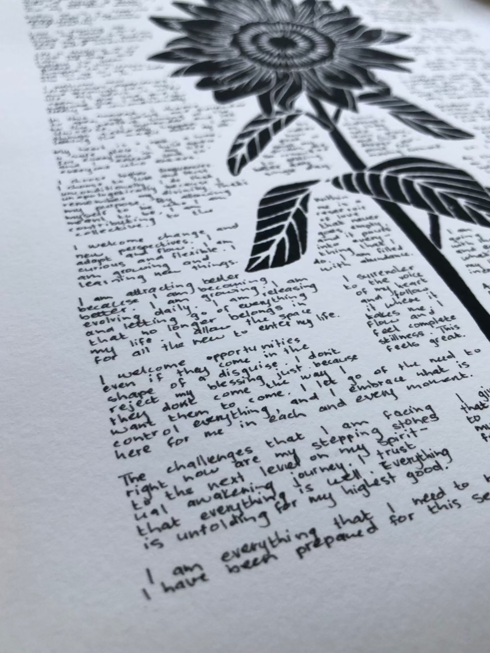 Close up of a lino print of a sunflower surrounded by tiny handwritten positive affirmations photographed from the bottom corner of the print.