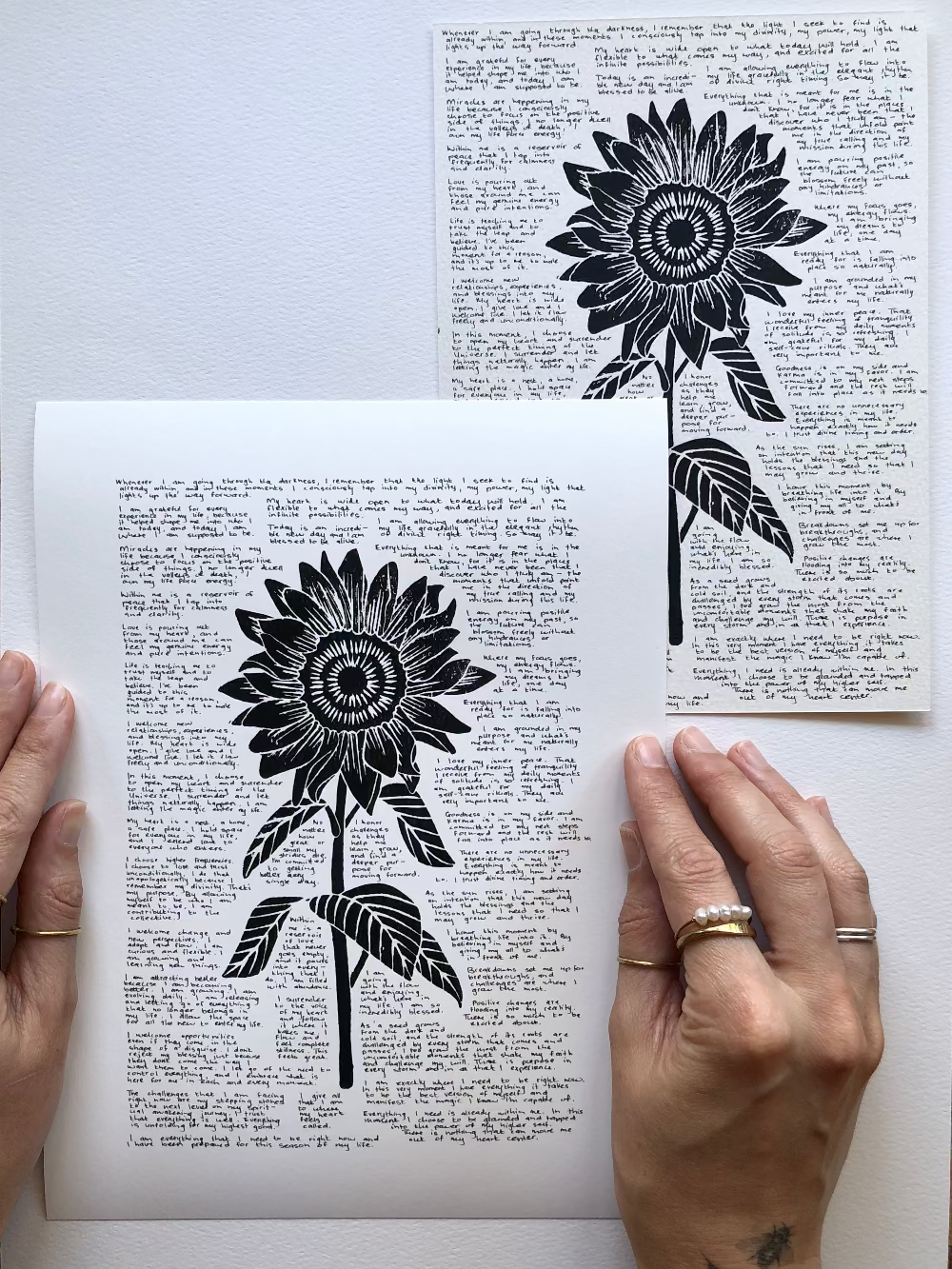Lino print of a sunflower surrounded by tiny handwritten positive affirmations photographed flat on a table next to the original artwork.