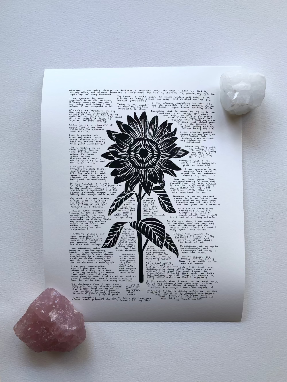 Lino print of a sunflower surrounded by tiny handwritten positive affirmations photographed flat on a table and held down with two crystals.