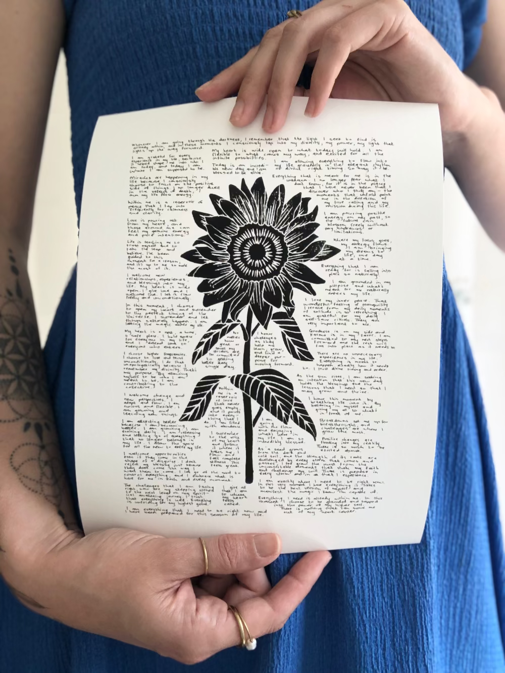 Lino print of a sunflower surrounded by tiny handwritten positive affirmations photographed being held by the artist.