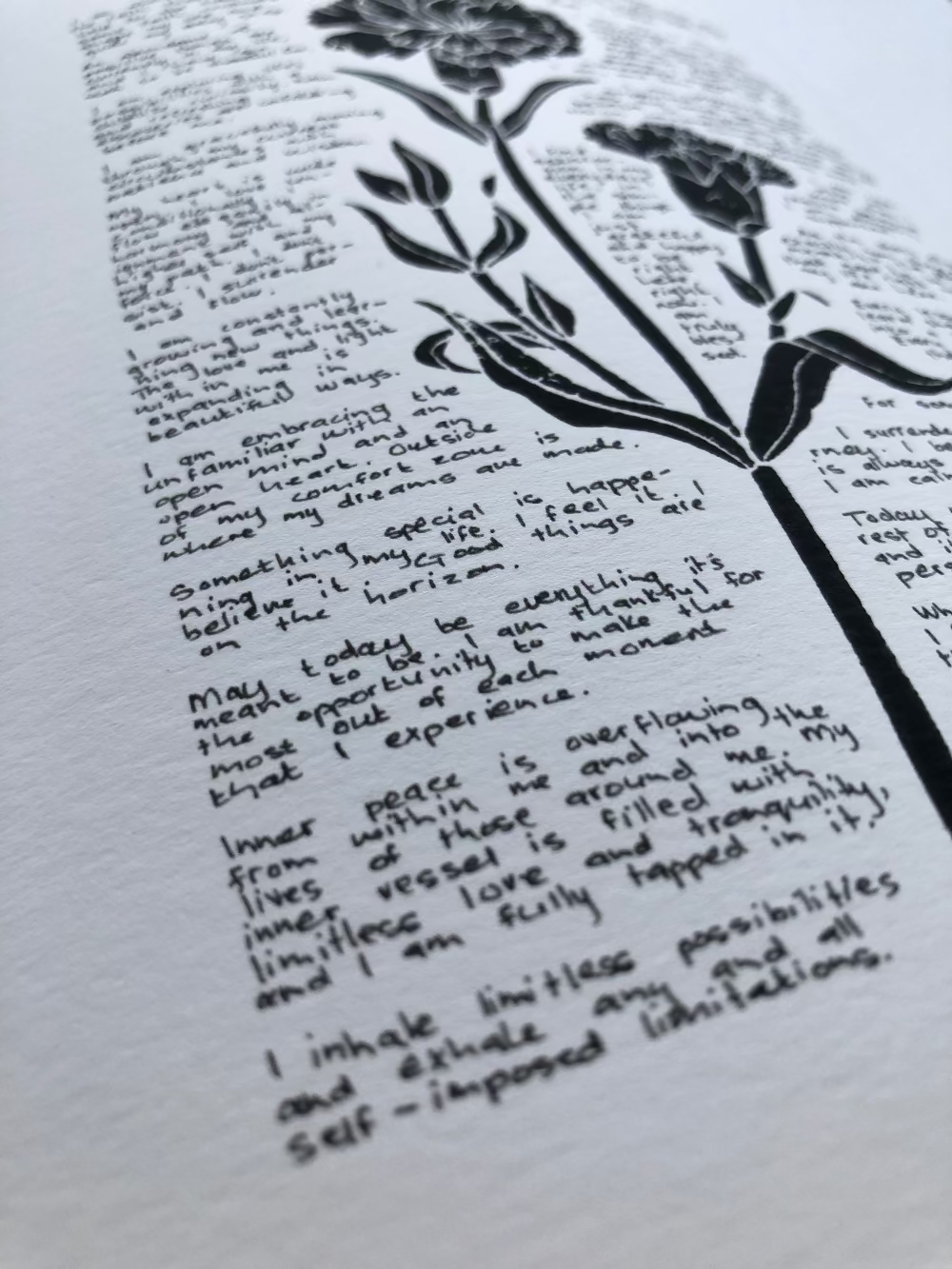 Close up of a lino print of a carnation flower surrounded by tiny handwritten positive affirmations, photographed from the corner of the print.