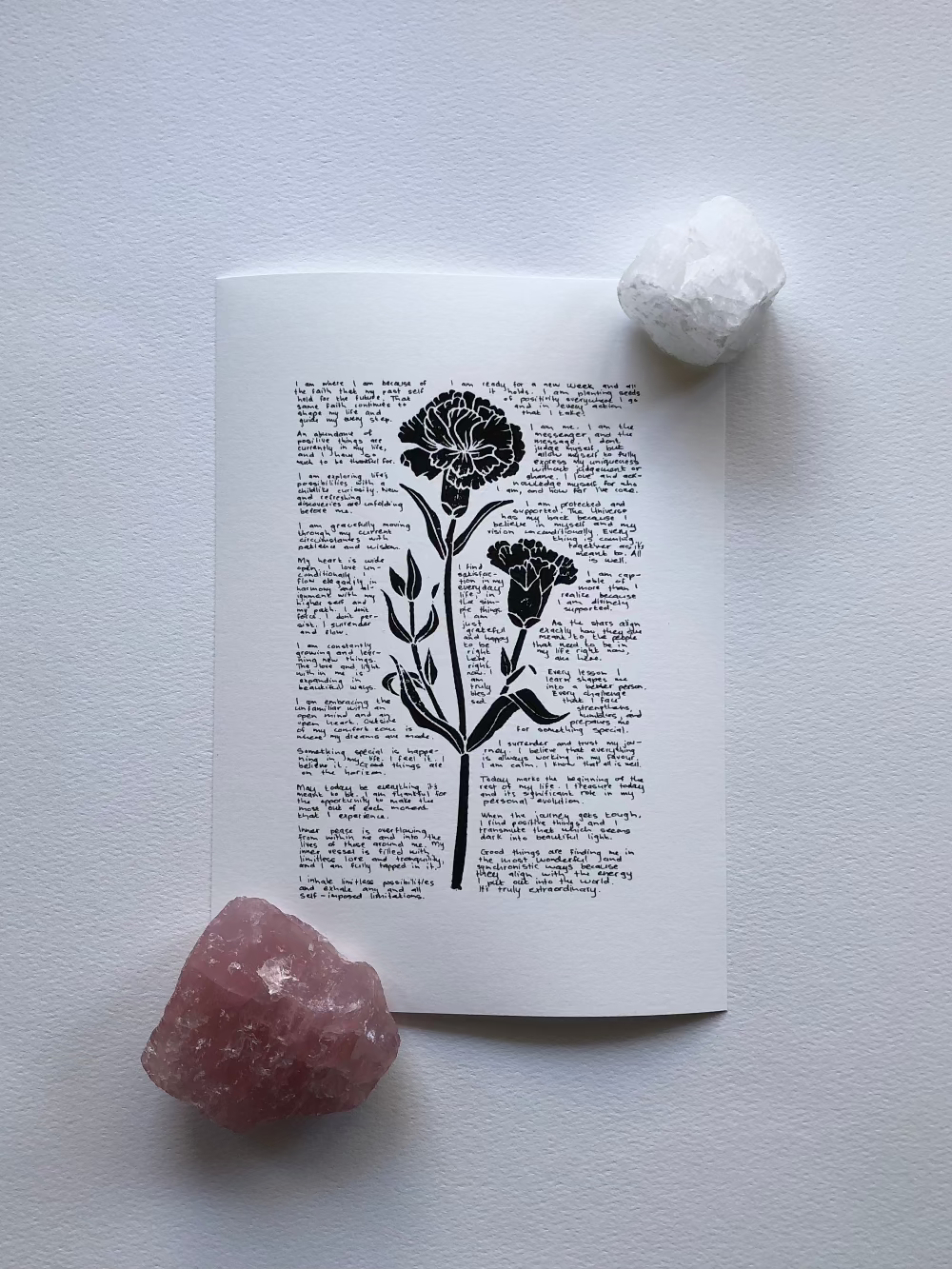 A lino print of a carnation flower surrounded by tiny handwritten positive affirmations is photographed flat on a table and held down with two crystals.