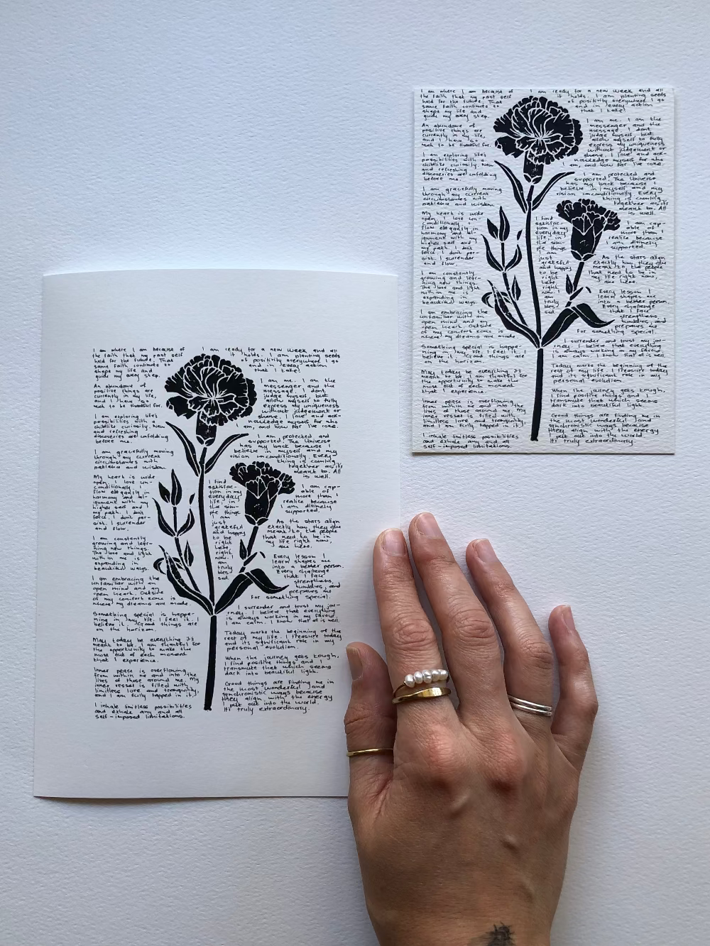 A lino print of a carnation flower surrounded by tiny handwritten positive affirmations, photographed flat on a table next to the original artwork.
