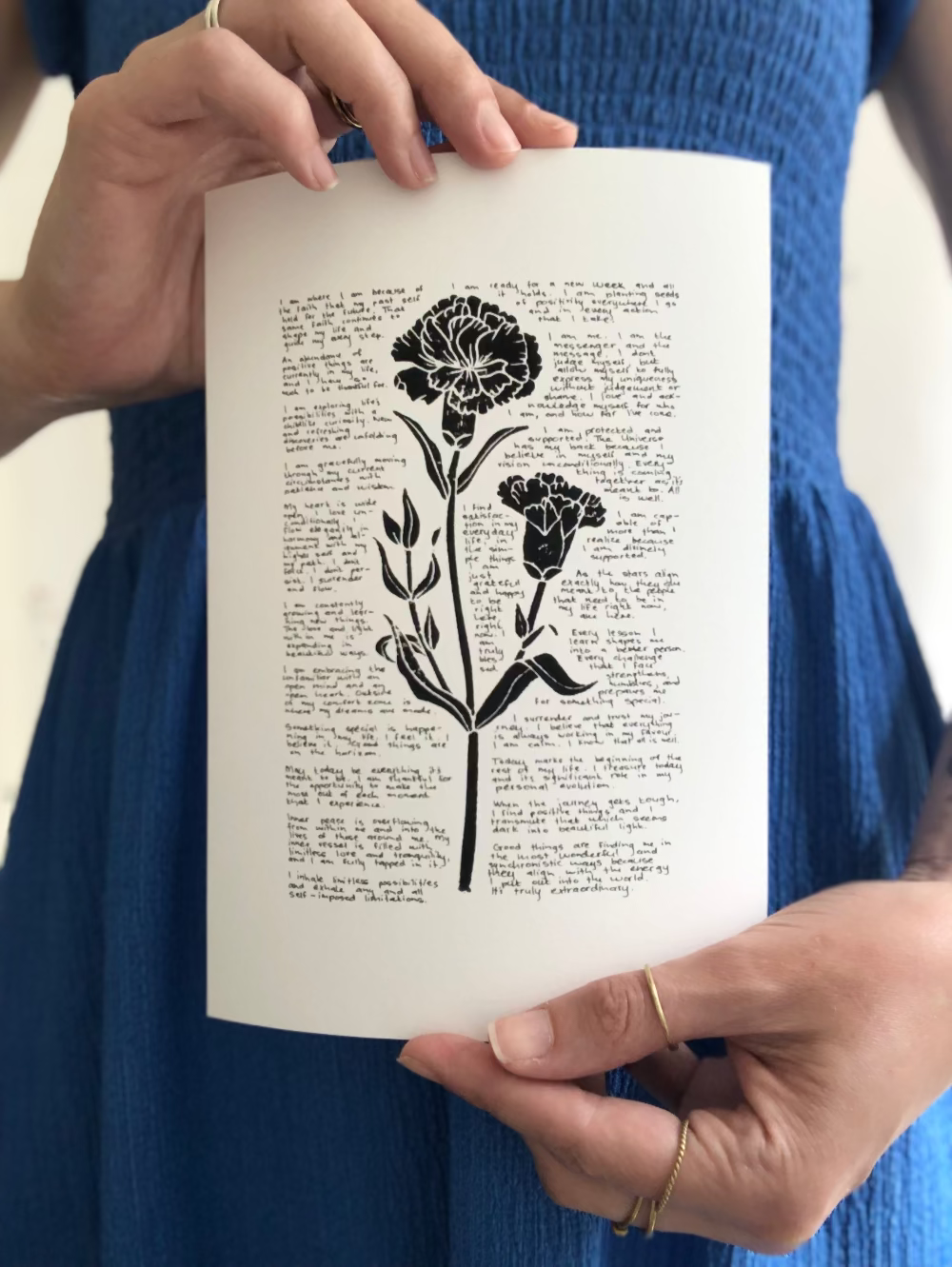 A lino print of a carnation flower surrounded by tiny handwritten positive affirmations, photographed being held by the artist