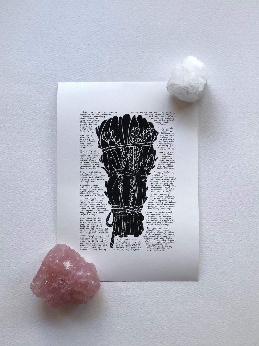 Black lino print surrounded with tiny handwritten positive affirmations photographed flat on a table and held down with two crystals.