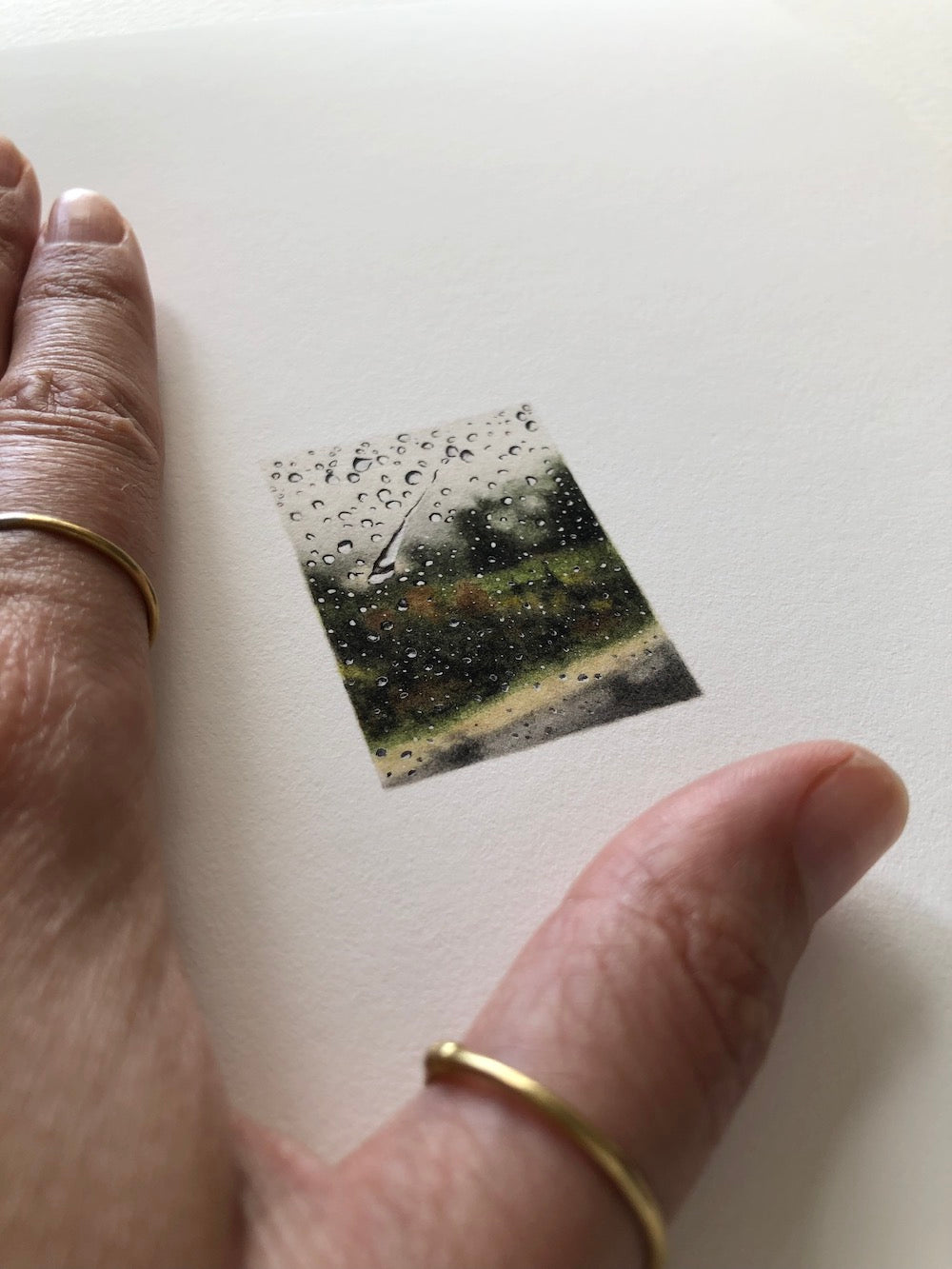 A hand resting on a piece of paper with a tiny print of a blurry landscape with raindrops against a window